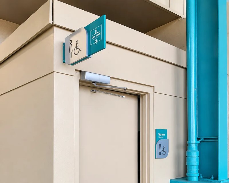 Rótulo de baño accesible en bandera sobre una puerta y placa junto a ella
